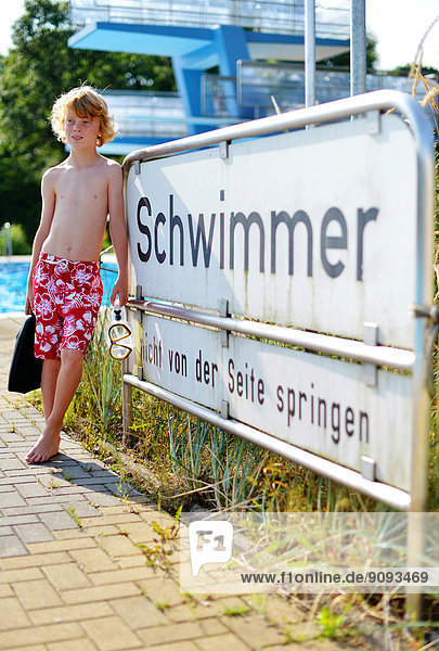 A boy with flippers and diving goggles is standing near the sign swimmers in an open air bath.