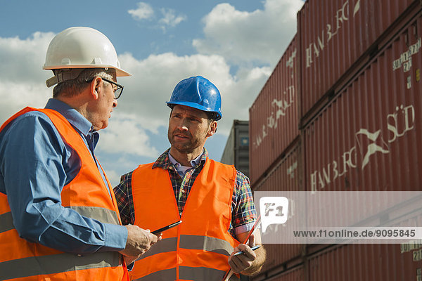 Two men with safety helmets and reflective vests talking at container port