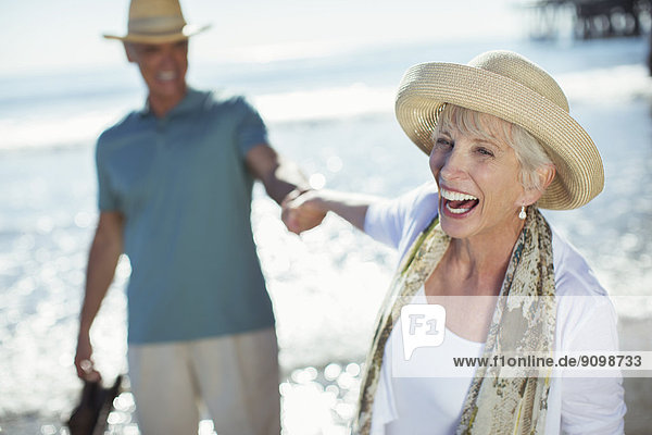 Enthusiastisches Seniorenpaar hält Hände am sonnigen Strand