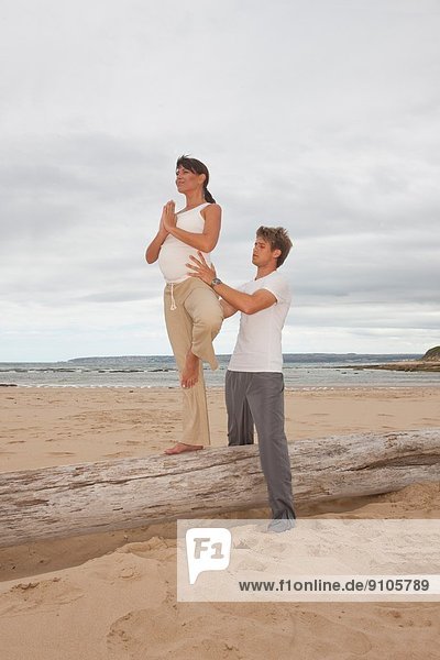 Schwangere mittlere erwachsene Frau mit Personal Trainerin  die Yoga am Strand praktiziert.