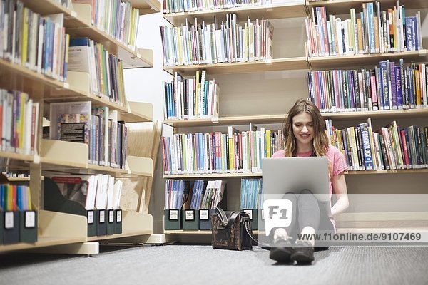 Junge Frau sitzt auf dem Boden in der Bibliothek mit Laptop
