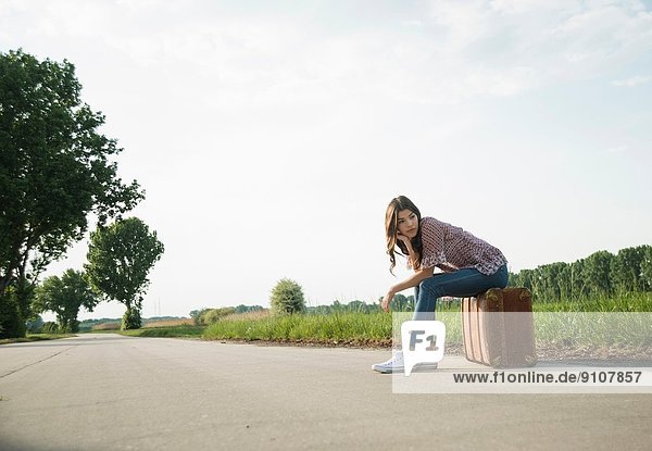 Young woman sitting on suitcase on country road