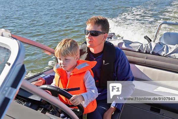 Young boy sitting on fathers lap steering motor boat