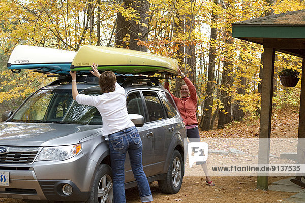 Two young women load kayaks on roof of vehicle at cottage near Oxtongue Lake  Muskoka  Ontario  Canada.