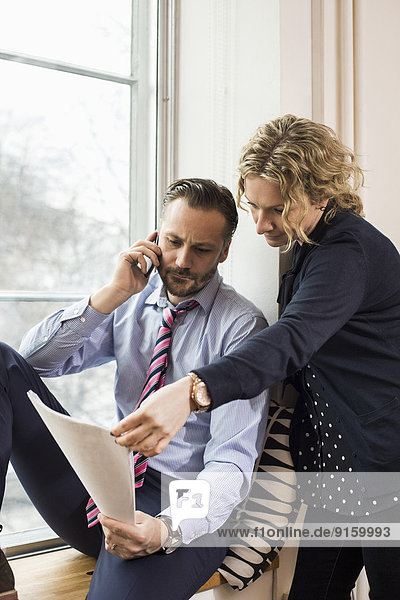 Businessman answering smart phone while discussing over document with female colleague by office window