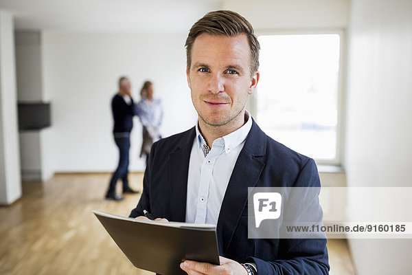 Portrait of confident real estate agent with couple standing in background at home