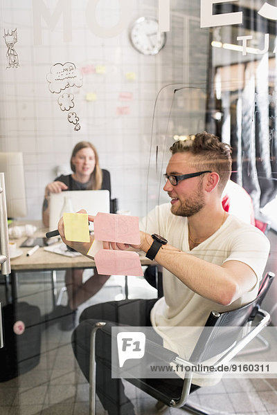 Young businessman writing ideas on adhesive notes with female colleague in background at new office
