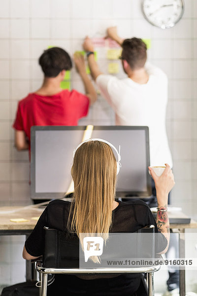 Rückansicht der jungen Geschäftsfrau beim Kaffee mit männlichen Kollegen im Hintergrund im Kreativbüro