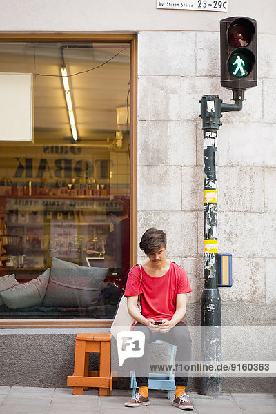 Volle Länge des jungen Mannes  der die SMS auf dem Handy auf dem Bürgersteig liest.