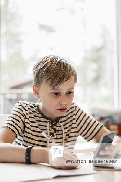 Junge mit Smartphone beim Schreiben am Tisch im Haus
