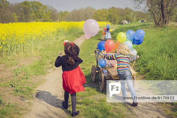 Drei Kinder mit Holzwagen und Luftballons unterwegs