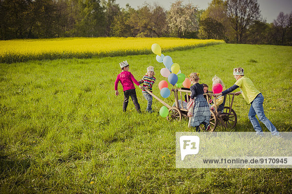 Sechs Kinder unterwegs mit Holzwagen und Luftballons