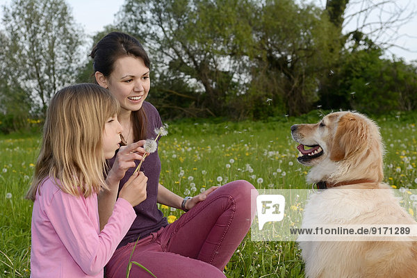 Young woman and little girl sitting with Golden Retriever on meadow