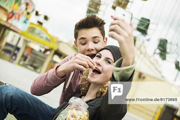 Portrait of teenage couple with popcorn photographing themself at fun fair