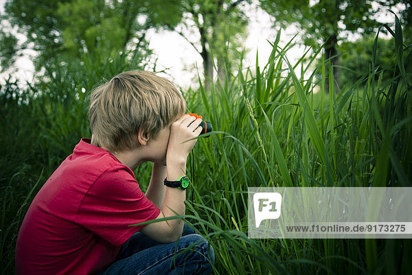 Kleiner Junge sitzt auf der Wiese und beobachtet etwas mit einem Fernglas.