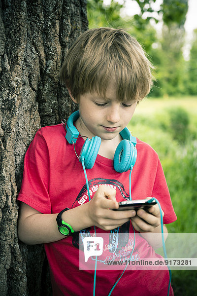 Portrait des Jungen mit Smartphone und Kopfhörer auf Baum gelehnt