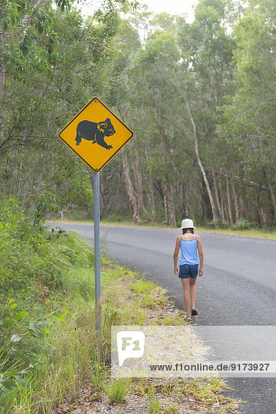 Australien  New South Wales  Pottsville  Straßenschild mit einem Koala-Bären und einem Mädchen  die im Pottsville Environmental Park entlang der Straße spazieren gehen.