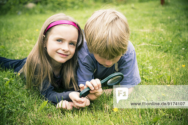 Bruder und Schwester liegen auf der Wiese und beobachten die Blume mit Lupe.