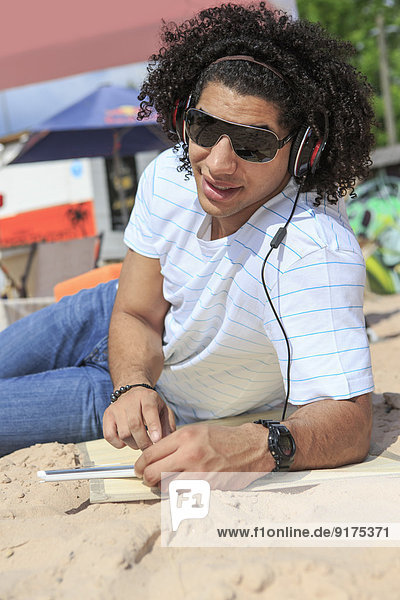 Porträt eines jungen Mannes mit digitalem Tablett beim Musikhören am Strand