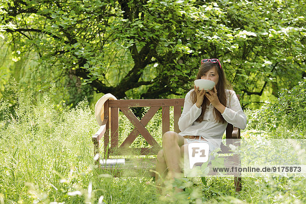 Young woman drinking on wooden bench in a garden