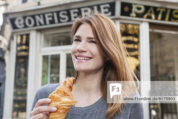 France  Paris  portrait of happy young woman with croissant in front of pastry shop