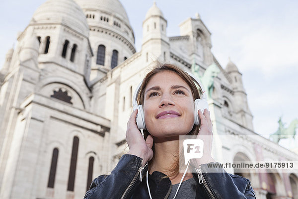 Frankreich  Paris  Porträt einer glücklichen jungen Frau  die Musik mit Kopfhörern vor Sacre Coeur hört.