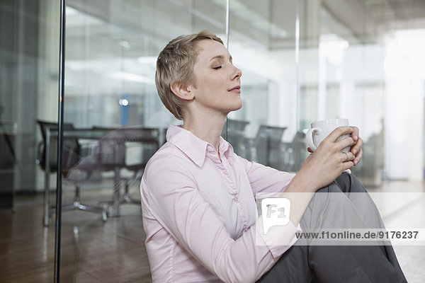 Deutschland  München  Geschäftsfrau im Büro  sitzend auf dem Boden mit Kaffeetasse