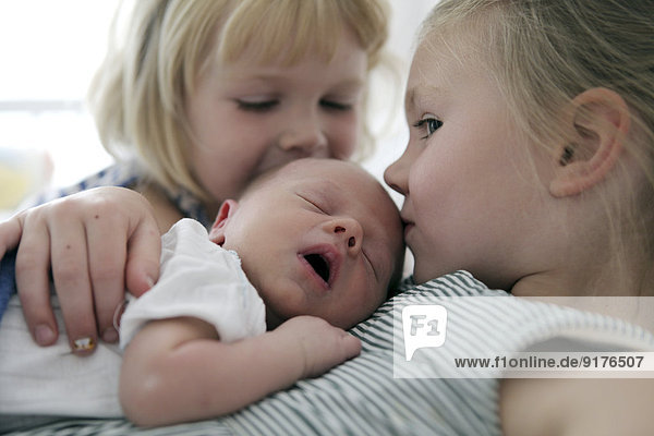 Zwei kleine Mädchen  die ihren neugeborenen Bruder küssen.