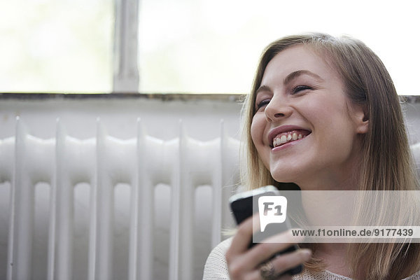 Lachende junge Frau mit Handy am Heizkörper