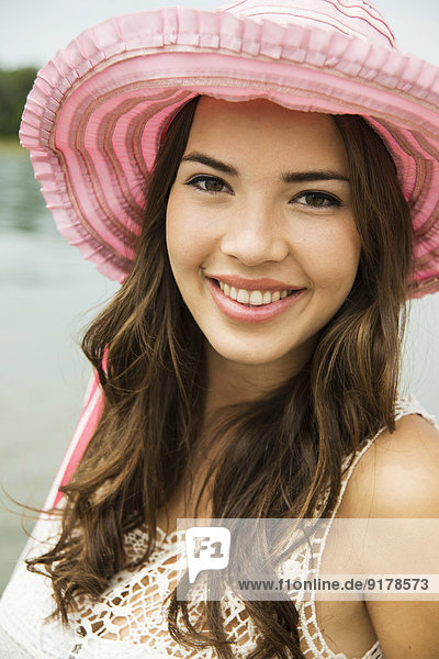 Porträt einer lächelnden jungen Frau am Strand mit rosa Sommerhut