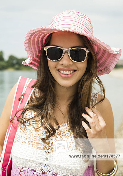 Porträt einer lächelnden jungen Frau mit Sonnenbrille und rosa Hut am Strand