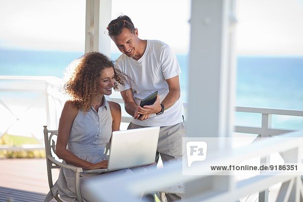 Paar mit Blick auf digitales Tablett und Laptop auf dem Balkon des Strandhauses