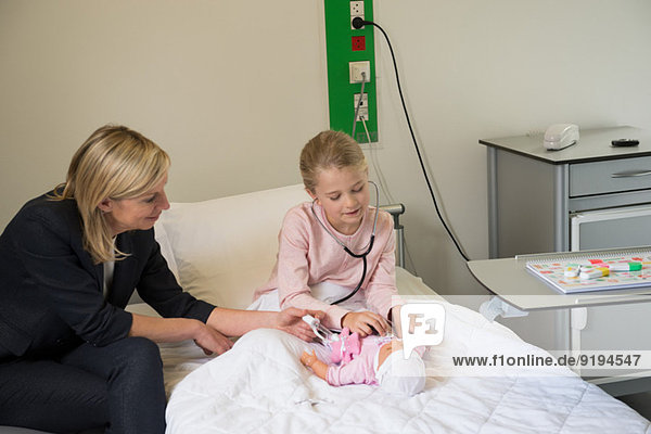 Mädchen untersucht eine Puppe mit Stethoskop und ihre Mutter sitzt neben ihr im Krankenhaus.