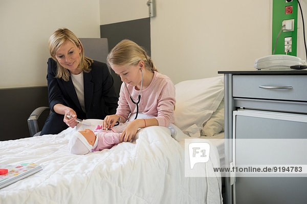 Girl examining a doll with stethoscope and her mother sitting beside her in hospital