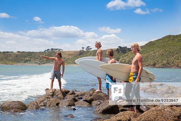 Four young male surfer friends pointing to sea from rocks
