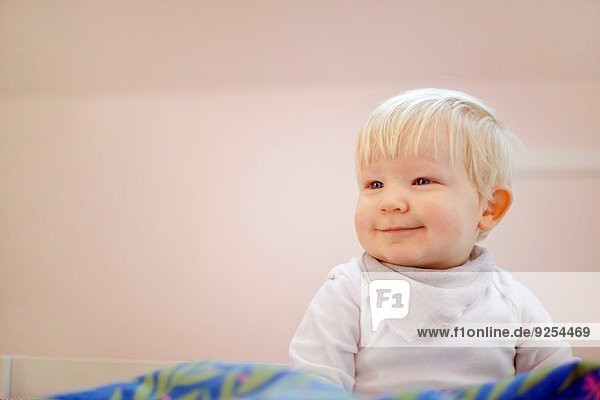 Porträt eines süßen vierzehn Monate alten Jungen der auf einer Bettdecke sitzt.