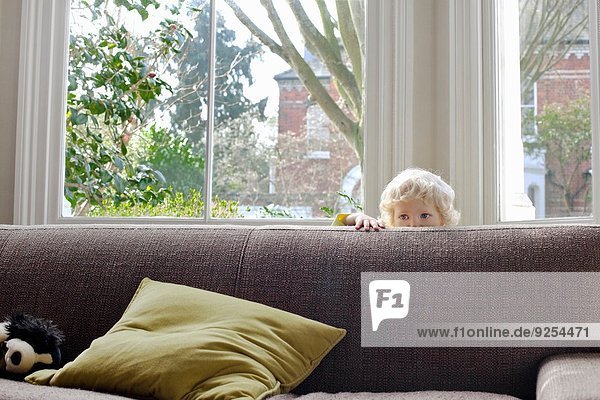 Porträt eines süßen blonden Kleinkindes  das sich hinter dem Sofa versteckt.
