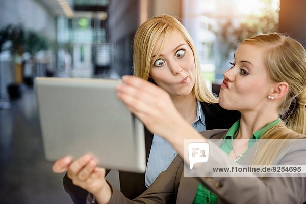 Zwei junge Geschäftsfrauen ziehen Gesichter und nehmen Selfie auf digitalem Tablett im Büro mit.