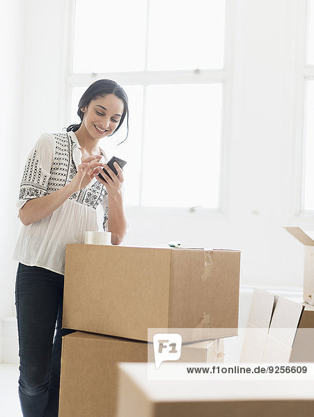 Young woman standing among boxes in new home and using mobile phone