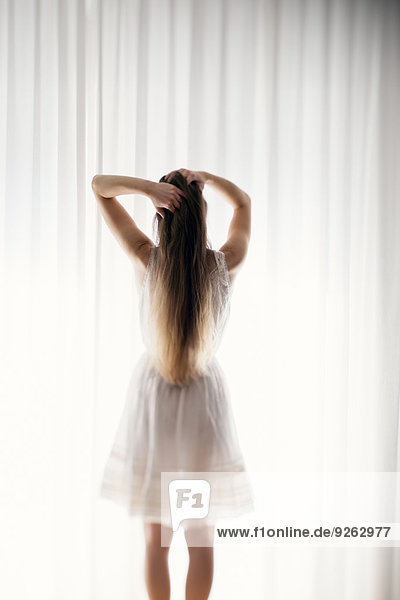 Junge Frau mit den Händen im Haar vor einem weißen Vorhang stehend  Rückansicht