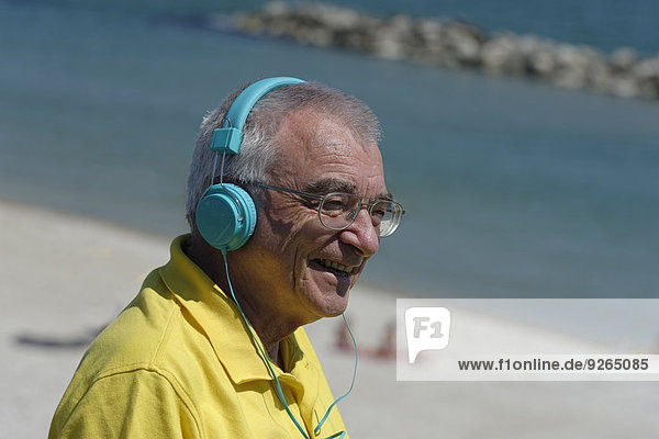 Porträt eines lächelnden älteren Mannes mit Kopfhörer vor einem Strand