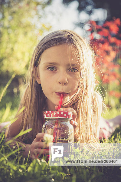 Porträt eines kleinen Mädchens  das auf einer Wiese im Garten liegt und Saft trinkt.