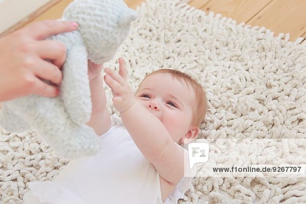 Weibliche Hand hält Teddybär für kleines Mädchen  das auf einem Teppich liegt