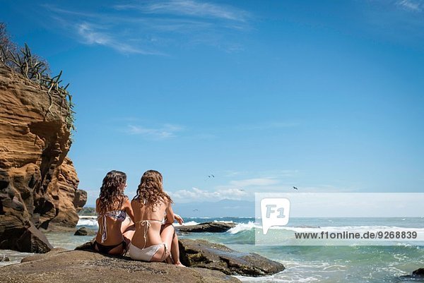 Zwei junge Frauen  die auf Felsen sitzen und auf den Ozean blicken.