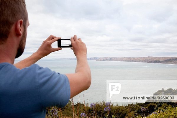 Rückansicht eines jungen Mannes  der mit einem Smartphone das Meer fotografiert.