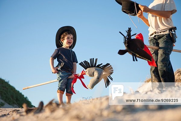 Zwei Brüder verkleidet als Cowboys mit Hobbypferden im Sand