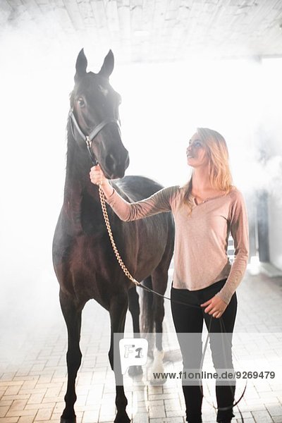 Young woman standing in stables with black horse