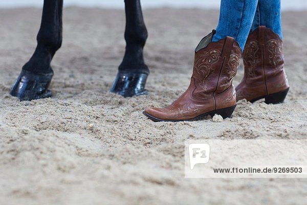 Cropped shot of horse hooves and young woman in cowboy boots