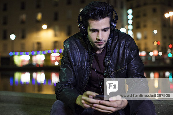 Junger Mann sitzt nachts allein im Freien und hört Musik mit Kopfhörern.