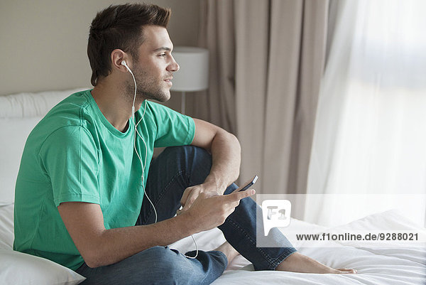 Junger Mann sitzt auf dem Bett und hört Musik mit dem Smartphone.
